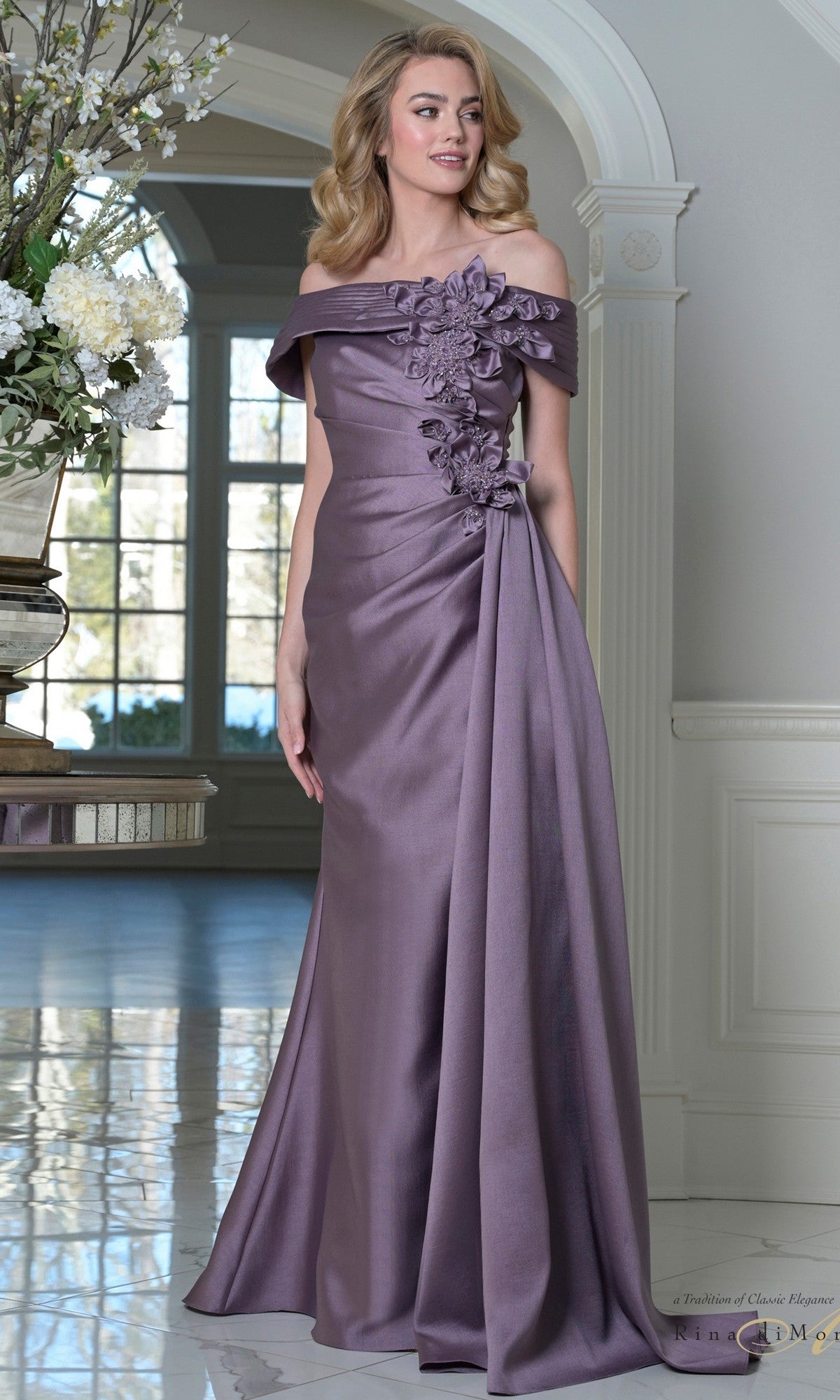 A woman stands indoors in the Rina Di Montella RD3528 long formal dress in Victorian Lilac, featuring an off-shoulder neckline and 3D floral appliques on the bodice. Marble flooring and decorative flowers are seen in the background.